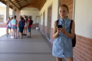 <colegiala-parada-en-el-patio-de-la-escuela-primaria-usando-un-telefono-inteligente>
